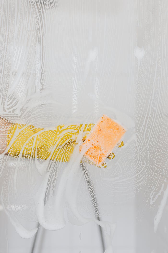 A gloved hand cleans glass with a sponge and soapy water, ensuring hygiene.